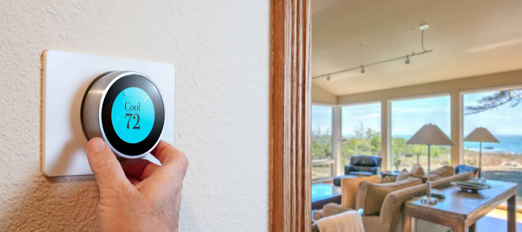 hand reaching up to adjust a smart thermostat with a living room setting at the beach in the background