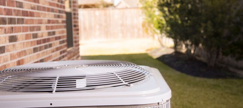 A close-up of an outdoor air conditioning unit next to a brick house, with a fenced backyard and green grass in the background on a sunny day.