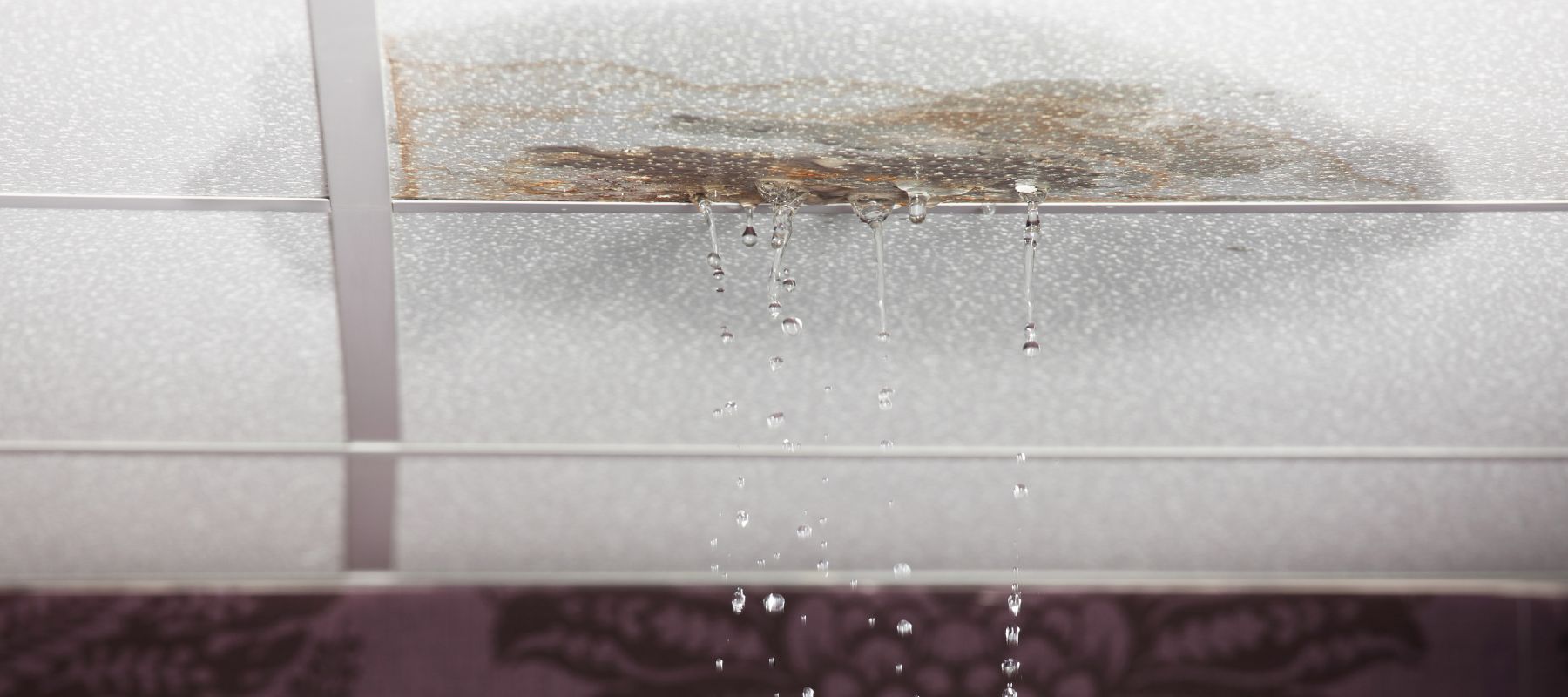 Water leaking through a stained ceiling tile, with several streams of water dripping down and visible damage around the leak.